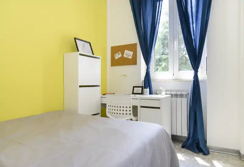 Dormitorio luminoso y ordenado con cama en primer plano, escritorio blanco junto a la ventana y cortinas azul oscuro que enmarcan la luz natural.