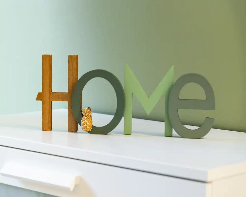 Gros plan décoratif de lettres en bois "HOME" posées sur une commode blanche devant un mur vert pâle — un détail de mise en scène qui apporte chaleur et caractère.