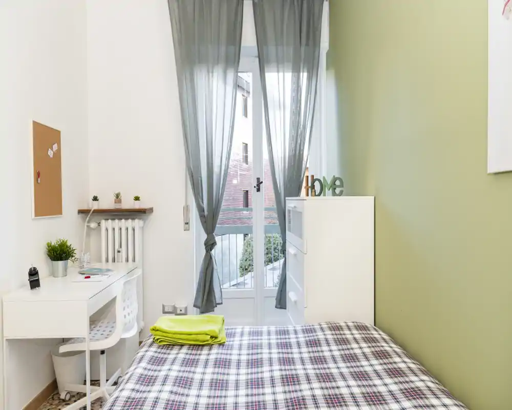 Chambre simple lumineuse et bien aménagée avec lit à motif, petit bureau et porte donnant sur le balcon qui apporte la lumière naturelle — idéale pour étudiants ou courts séjours.
