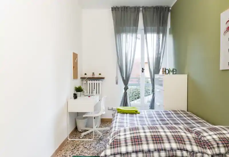 Chambre simple lumineuse et soignée avec une couette à carreaux, un petit bureau et une commode près d'une porte-fenêtre laissant entrer la lumière naturelle.
