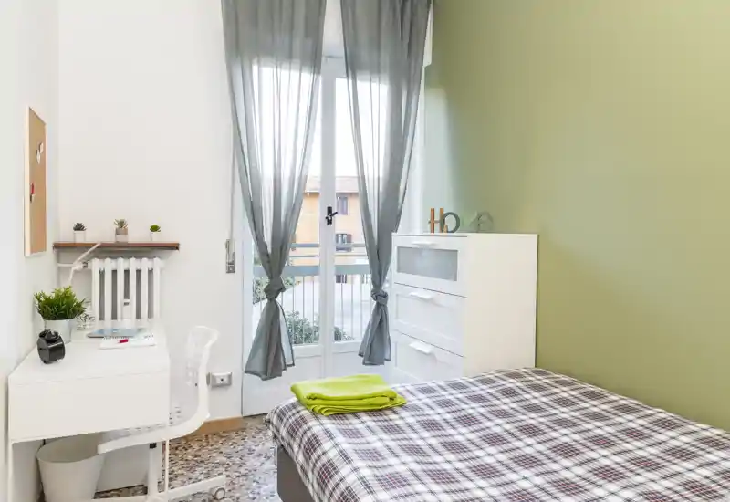Chambre simple lumineuse et bien aménagée avec couette à carreaux, lumière naturelle depuis la porte-fenêtre, petit bureau et rangement — parfaite pour un étudiant ou une personne seule.