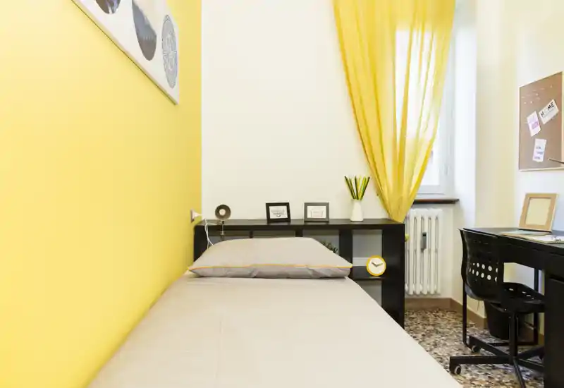 Chambre compacte et lumineuse avec lit simple, accent jaune chaleureux et coin bureau soigné. La lumière naturelle et la décoration sobre la rendent parfaite pour une location courte durée ou étudiante.