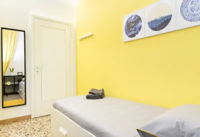 Chambre simple lumineuse et compacte avec lit contre un mur jaune vif, linge de lit soigné et décor mural — idéale pour un séjour confortable en solo.