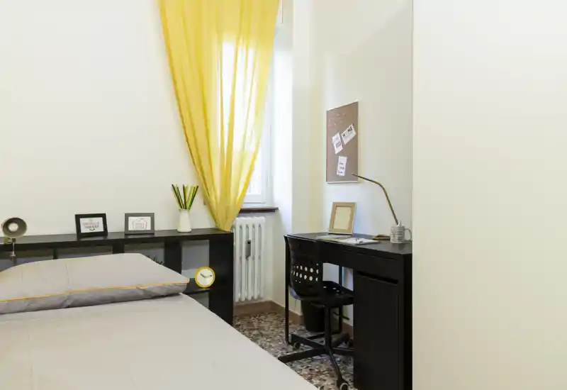 Chambre lumineuse et accueillante avec lit bien fait, étagère noire servant de tête de lit et un petit bureau près de la fenêtre encadrée d’un rideau jaune chaleureux.