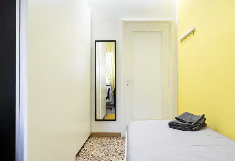 Chambre compacte avec un lit simple près d'un mur jaune, linge plié et un miroir pleine longueur reflétant une fenêtre voisine—lumineuse et minimaliste.