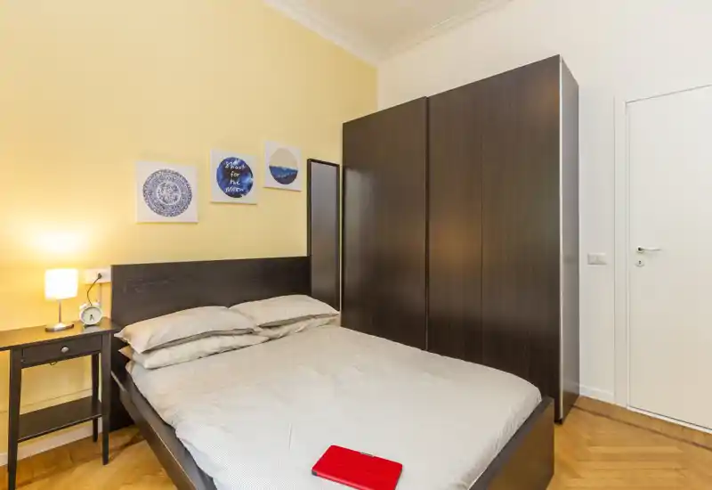 Sunny, well-styled bedroom with a double bed, bedside table and a large dark wardrobe; warm lighting and parquet flooring create a cozy atmosphere ideal for rental listings.