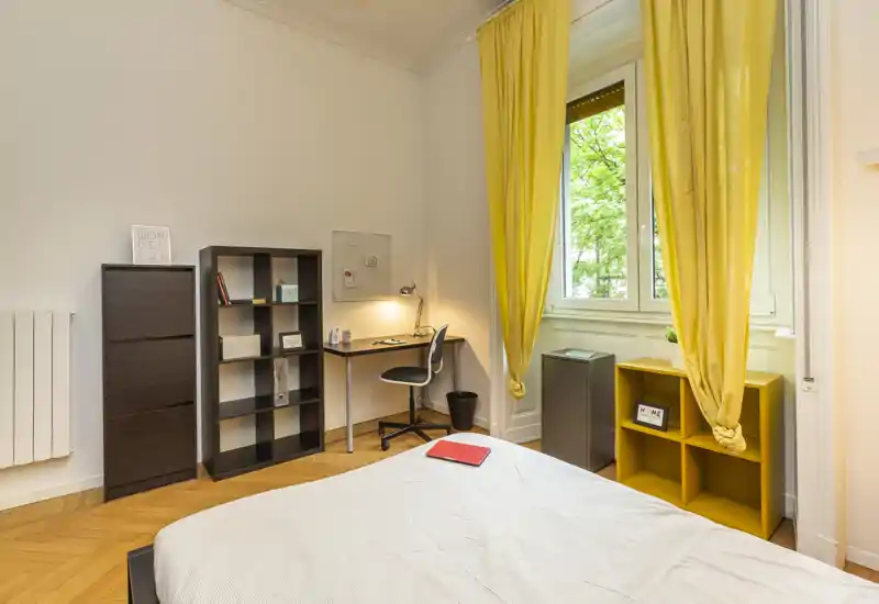 Bright, well-staged bedroom with a comfortable bed in the foreground, a desk and shelving for work or storage, and large window with yellow curtains bringing in natural light.