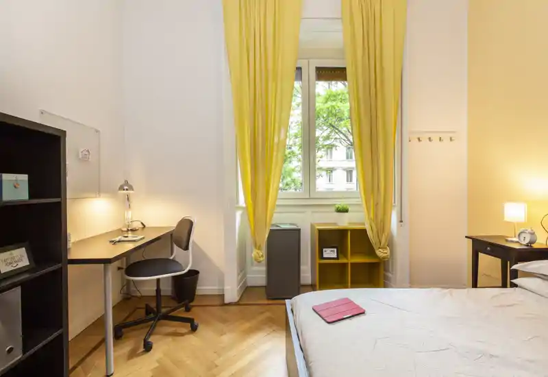 Bright, well-staged bedroom with a comfortable bed, warm bedside lighting and a small desk by the window framed by yellow curtains.