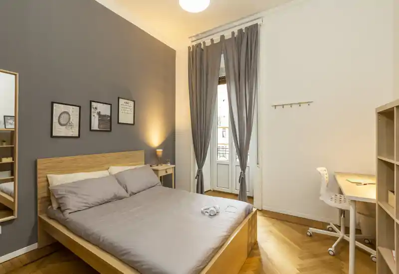 Camera da letto moderna e luminosa con letto matrimoniale in legno, comodino e una piccola postazione di lavoro vicino alla finestra. Parete grigia d'accento e tessili morbidi creano un’atmosfera accogliente perfetta per gli annunci.