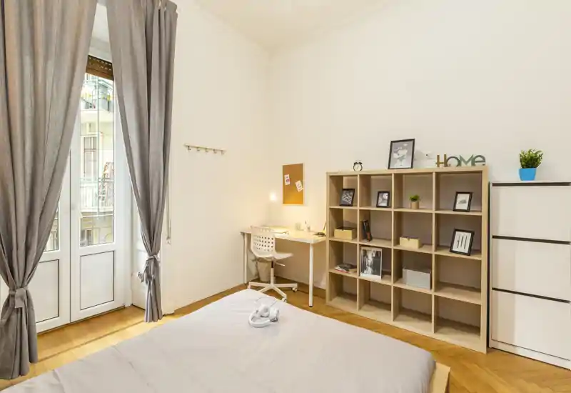 Camera da letto luminosa e ben arredata con letto ampio, angolo studio e libreria. La luce naturale dalle porte del balcone e i toni neutri creano un ambiente accogliente.