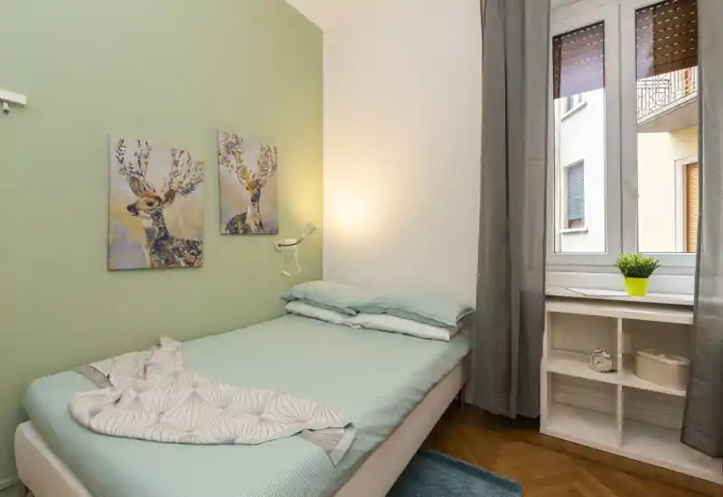 Dormitorio compacto y luminoso con cama individual vestida en tonos aqua, cuadros de ciervos, lámpara de lectura y ventana con una pequeña planta en la repisa — ordenado y acogedor para un huésped.