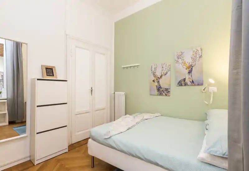 Dormitorio luminoso y cuidado con cama doble, pared verde y cuadros decorativos. La luz natural y artificial hace que la habitación sea acogedora y perfecta para un anuncio de alquiler.