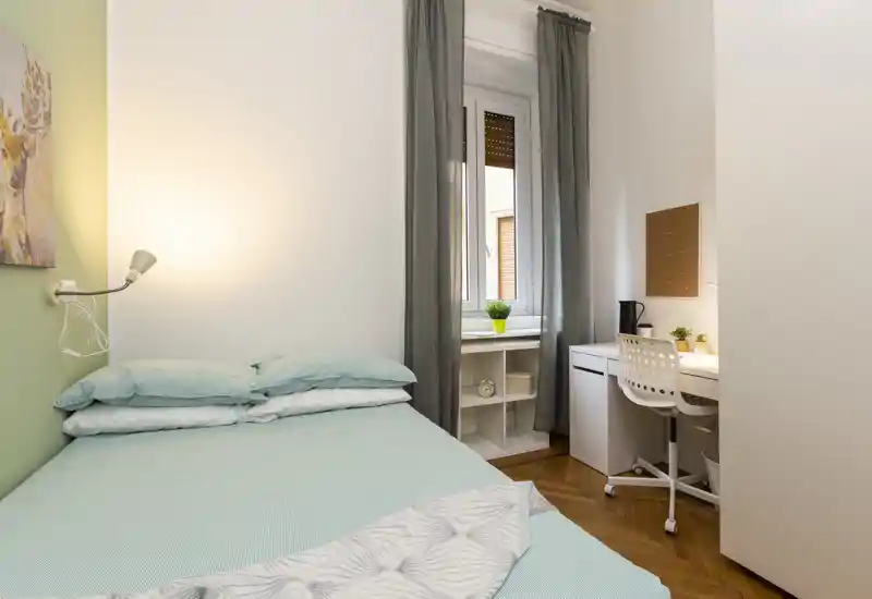 Dormitorio pequeño y luminoso con cama doble bien hecha en tonos pastel, lámpara de pared y un espacio de trabajo compacto junto a la ventana. Decoración neutra, cortinas grises y suelo de parquet para una atmósfera tranquila y acogedora.