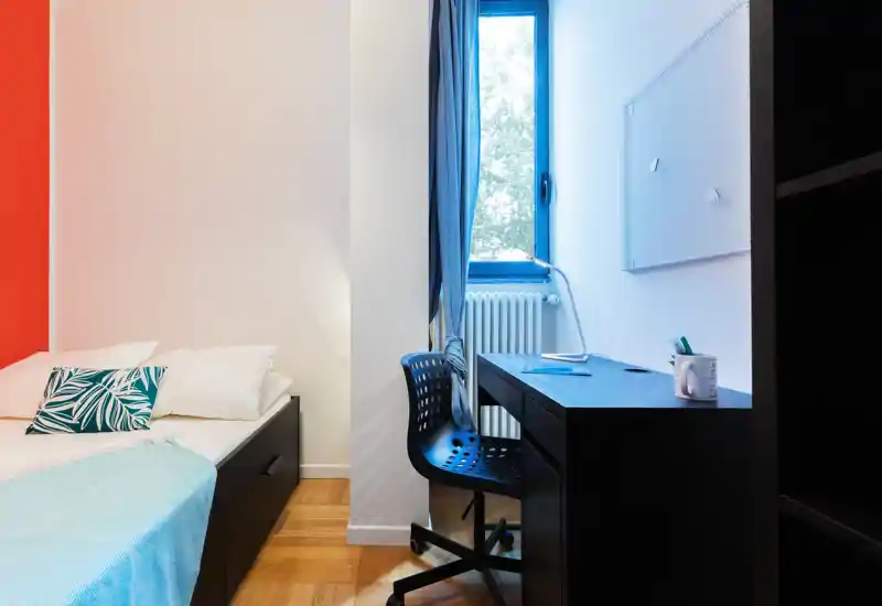 Dormitorio luminoso y ordenado con una cama individual cómoda, cojín decorativo y un escritorio funcional junto a la ventana — ideal para estudiar o trabajar desde casa.