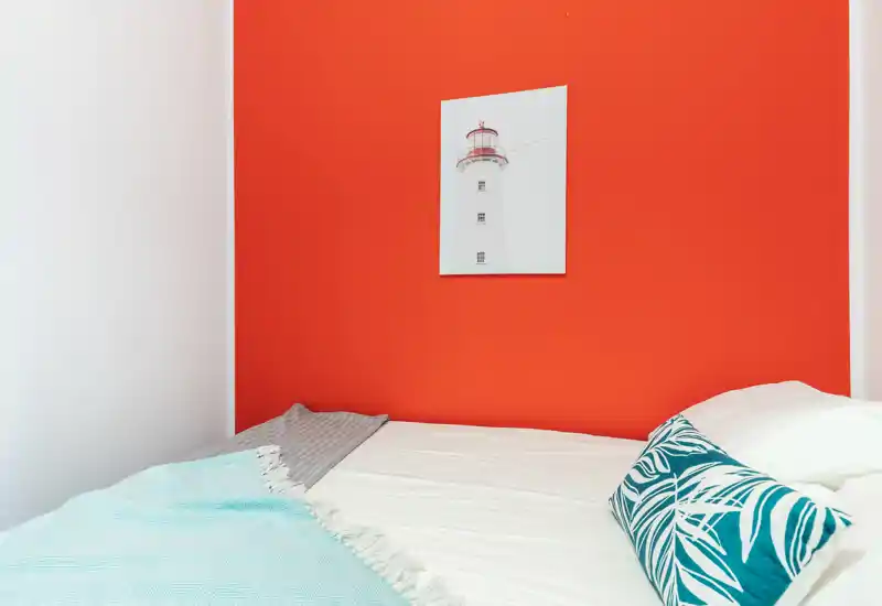 Rincón de dormitorio moderno y luminoso con cama blanca frente a una pared coral intensa, cojín decorativo y una manta ligera para mayor confort.