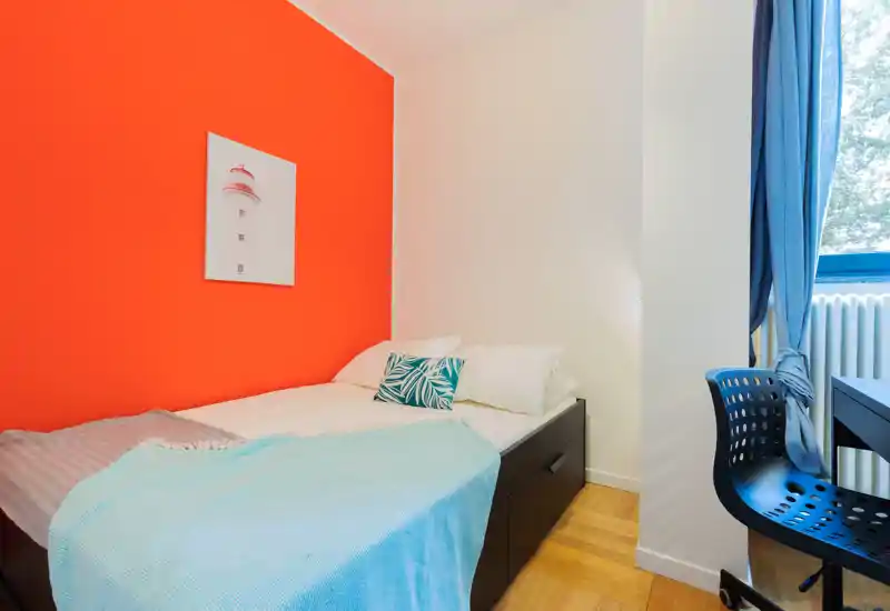 Dormitorio individual moderno y luminoso con una pared de acento naranja, cama ordenada con base de almacenamiento, cojín decorativo y manta azul clara, además de escritorio y silla junto a la ventana.