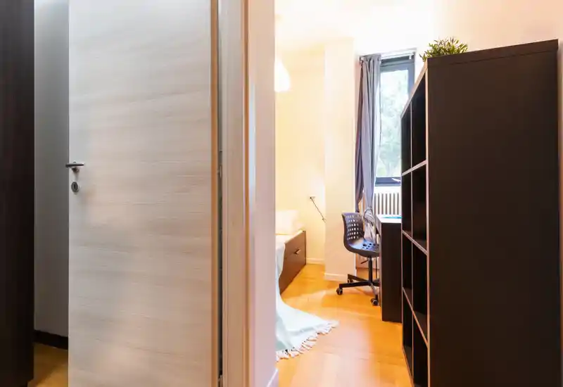 Pequeña habitación vista a través de una puerta abierta, con cama individual, mesita y un puesto de trabajo junto a la ventana. Espacio luminoso, mobiliario sencillo y suelo de madera cálido.