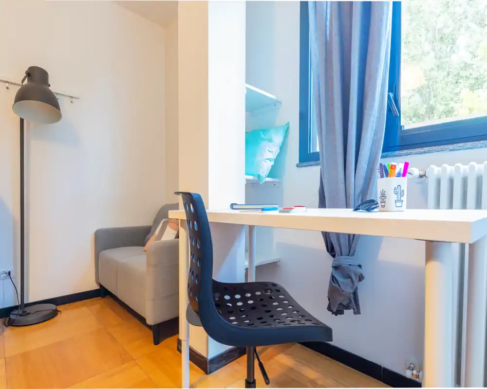 Rincón de sala luminoso y ordenado con un sofá compacto, lámpara de pie y un pequeño escritorio junto a la ventana — ideal para relajarse o trabajar.