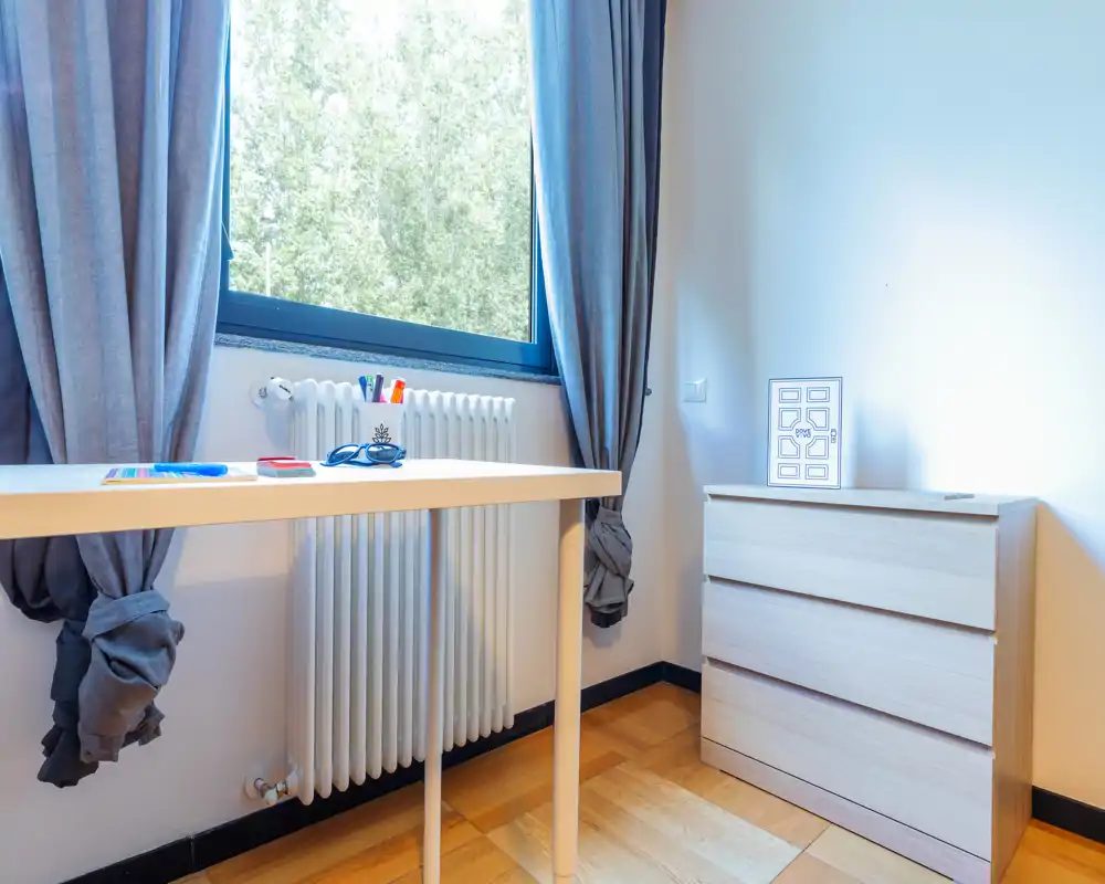 Habitación pequeña y luminosa con ventana, radiador, escritorio sencillo y cómoda de tres cajones; decoración neutra y luz natural, apta como dormitorio pequeño o despacho.