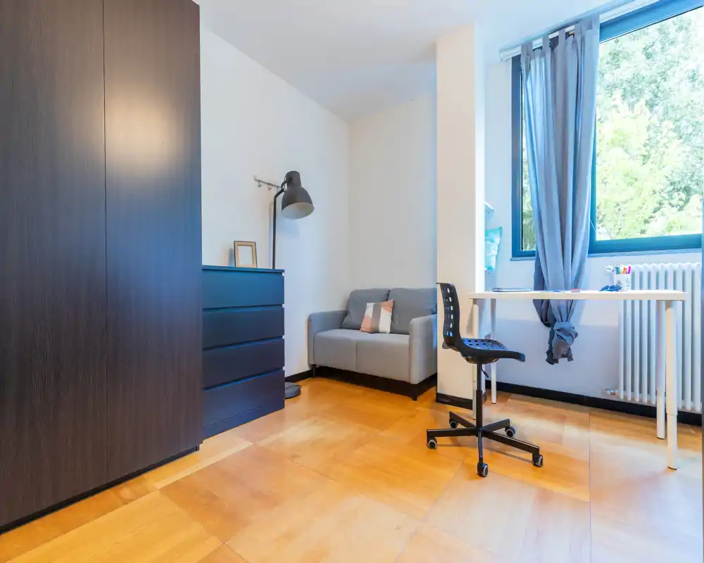 Rincón de dormitorio compacto con armario, cómoda, un pequeño sofá y escritorio junto a una gran ventana que proporciona abundante luz natural.