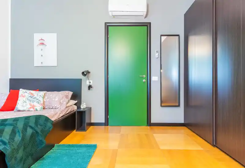 Dormitorio moderno y luminoso con cama doble visible, cojines coloridos y un gran armario oscuro. Suelo de madera cálida, puerta verde como acento e iluminación contemporánea que aporta calidez.