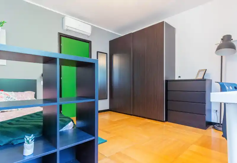 Dormitorio moderno y luminoso con cama visible, armario y cómoda de madera oscura, y una estantería en primer plano. Suelo de parquet cálido y ambiente cómodo y bien iluminado.