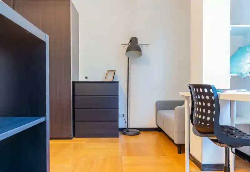 Área de estar luminosa y minimalista con sofá compacto, lámpara de pie y una cómoda pequeña — suelos de madera y tonos neutros crean un ambiente sereno, perfecto para descansar o trabajar.