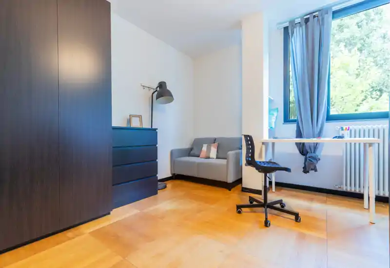 Rincón de dormitorio compacto con armario, cómoda, un pequeño sofá y escritorio junto a una gran ventana que proporciona abundante luz natural.