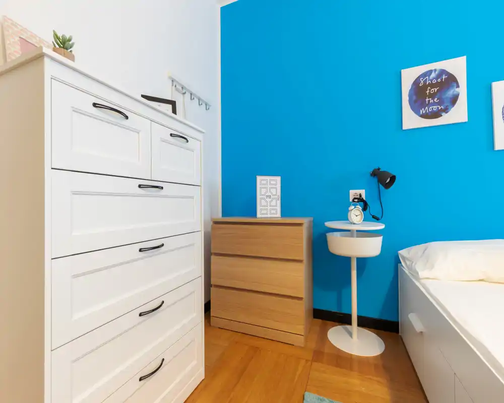 Rincón de dormitorio moderno y luminoso con cómoda blanca, mesita de madera y cama parcialmente visible frente a una pared azul intensa.