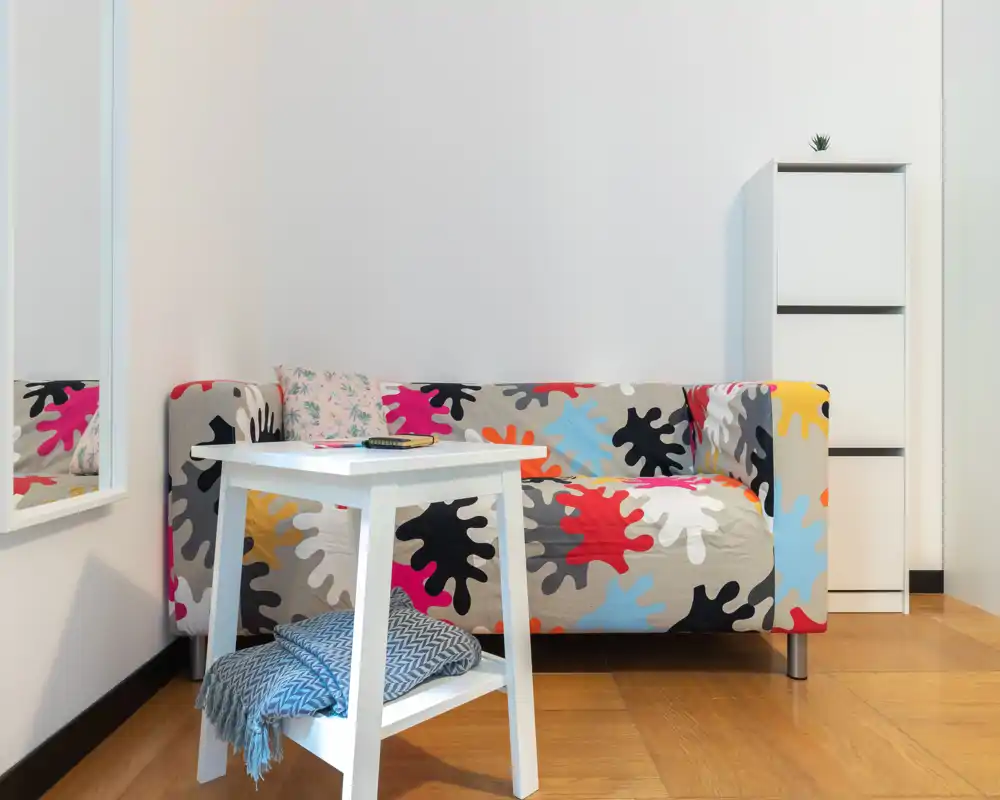 Rincón de estar moderno y luminoso con un sofá colorido de estampado, una mesita blanca y una manta decorativa — un espacio acogedor y bien iluminado para relajarse.