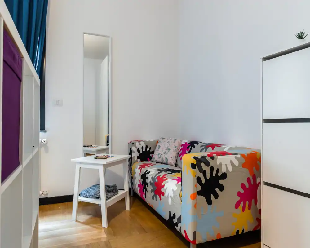 Pequeña zona de estar con sofá estampado y colorido, mesa auxiliar blanca y espejo alto: rincón luminoso, acogedor y con estilo para relajarse.