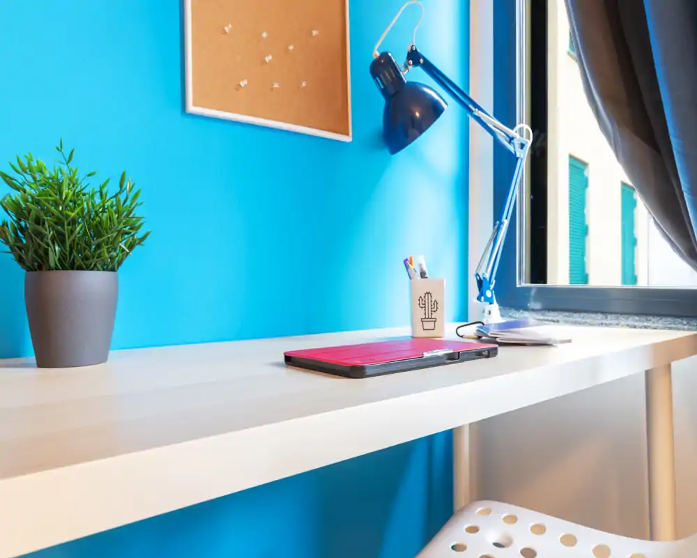 Rincón de escritorio moderno y luminoso con planta, lámpara y ventana — ideal como oficina en casa o área de estudio compacta.