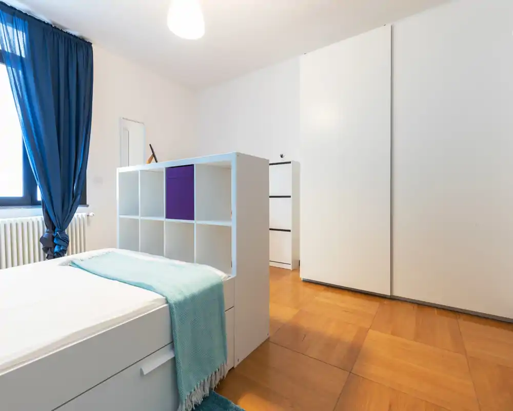Dormitorio moderno y luminoso con cama individual separada por una estantería baja, amplio armario blanco y cortinas azules que aportan un estilo fresco y minimalista.
