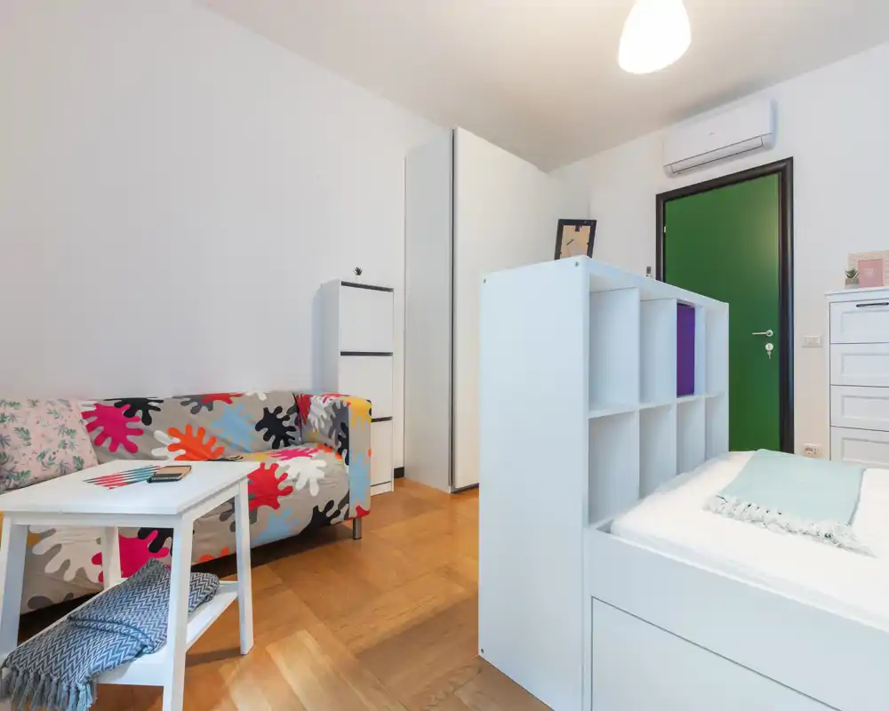 Dormitorio moderno y luminoso con una cama modular blanca en primer plano y un sofá colorido a la izquierda. Suelo de parquet cálido, muebles de almacenamiento sencillos y aire acondicionado sobre la puerta.
