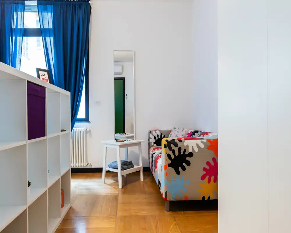 Zona de estar moderna y luminosa con un sofá de estampado colorido, una estantería blanca y una ventana con cortinas azul intenso que aportan un aire fresco y estiloso.