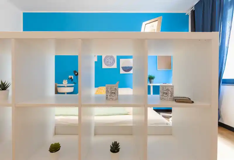 Dormitorio moderno visto a través de una estantería blanca de cubos: cama bien hecha, pared azul, cuadros y luz natural desde la ventana.