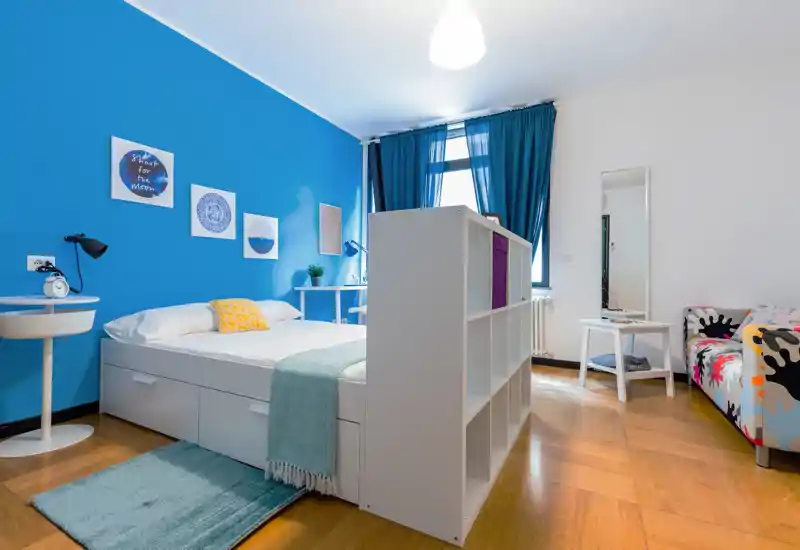 Dormitorio luminoso y con estilo con cama doble, cajones de almacenaje, pared azul de acento y estantería divisoria; buena luz natural y textiles acogedores.
