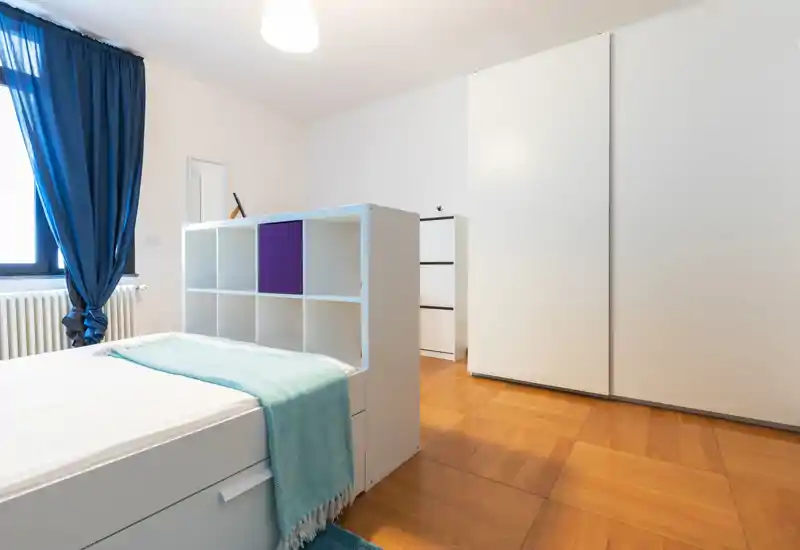 Dormitorio moderno y luminoso con cama individual separada por una estantería baja, amplio armario blanco y cortinas azules que aportan un estilo fresco y minimalista.