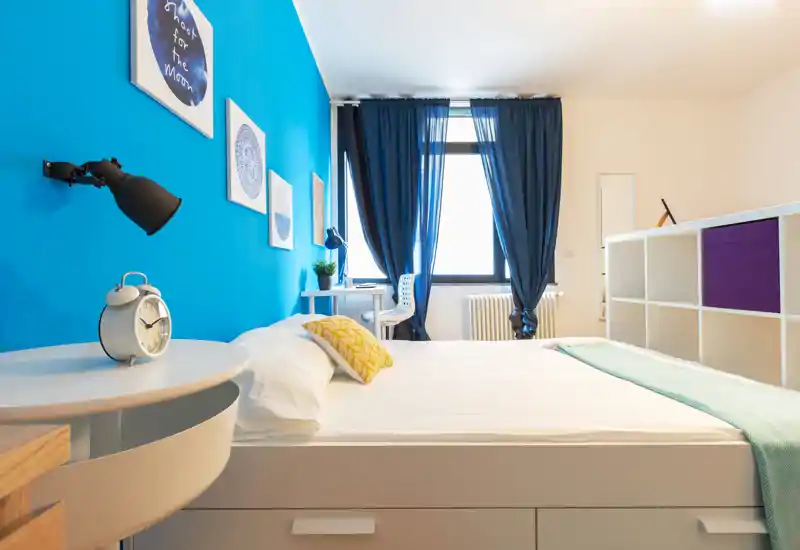 Dormitorio moderno y luminoso con una gran cama blanca, pared azul, escritorio junto a la ventana y estantería ordenada — fresco y elegante.