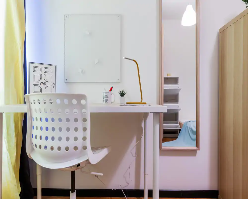 Rincón de dormitorio compacto con escritorio blanco y silla perforada; la cama sólo se ve parcialmente en el espejo alto a la derecha. Decoración minimalista y luminosa con lámpara de escritorio, planta pequeña y superficies ordenadas.