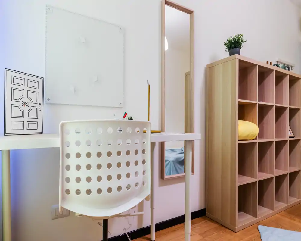 Esquina de dormitorio luminosa y ordenada con un pequeño escritorio y estantería; un espejo refleja parte de la cama y aporta profundidad.
