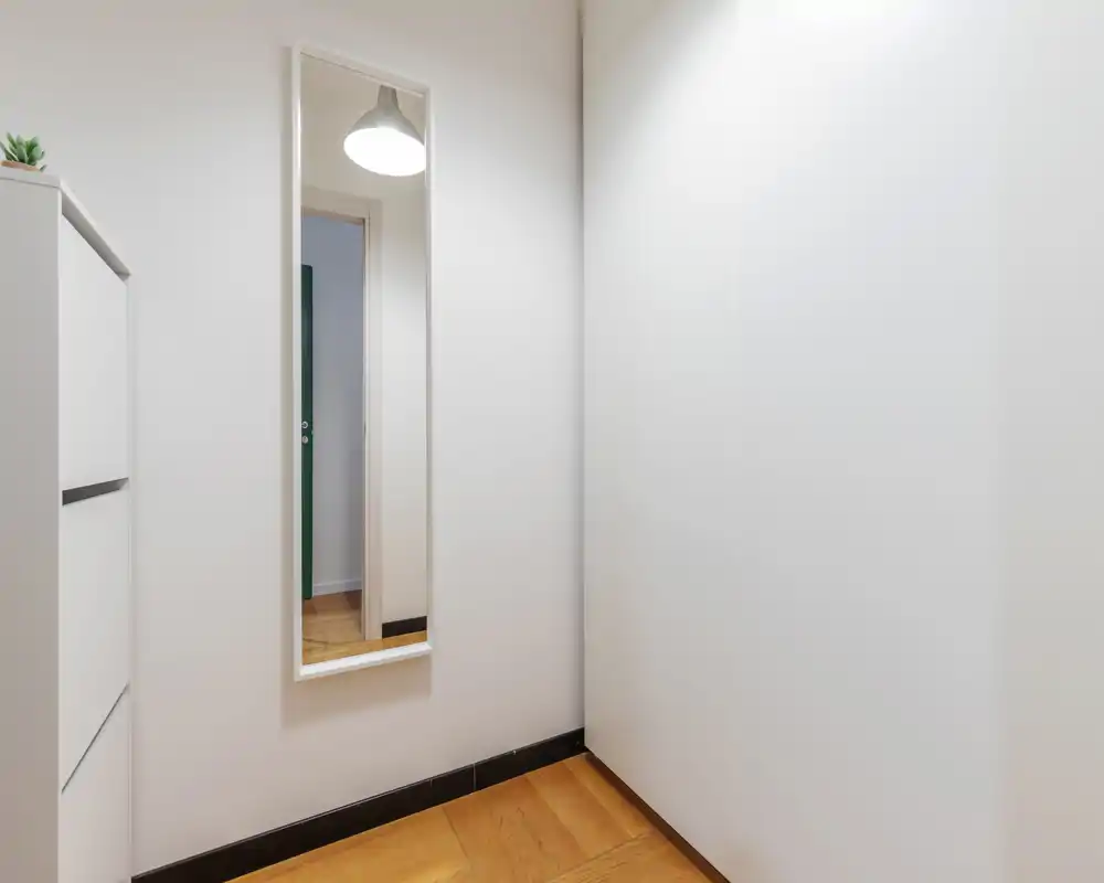 Small, minimal entrance or hallway corner with a full-length mirror and a narrow shoe/storage cabinet on warm wooden flooring.