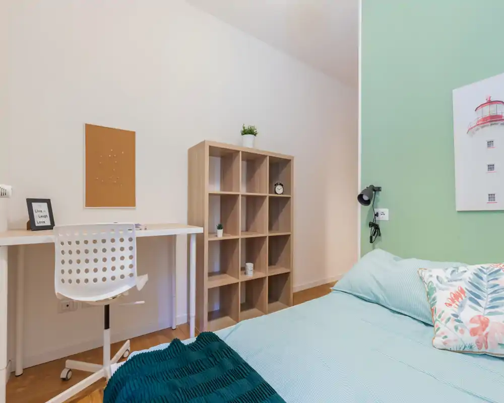 Bright, tidy bedroom with a visible double bed dressed in light linens, a bedside reading lamp and a small desk with chair — ideal for a student or professional.