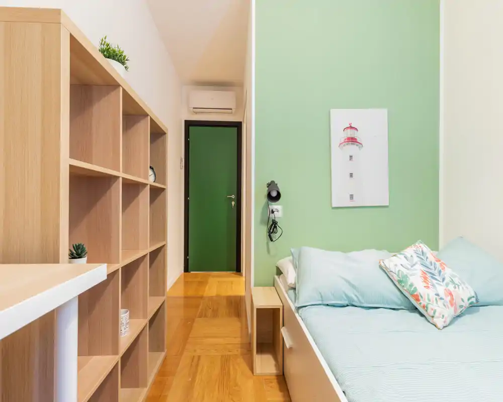 Bright, modern single bedroom with a neat bed, pastel green accent wall and wooden shelving providing storage and a cozy atmosphere.