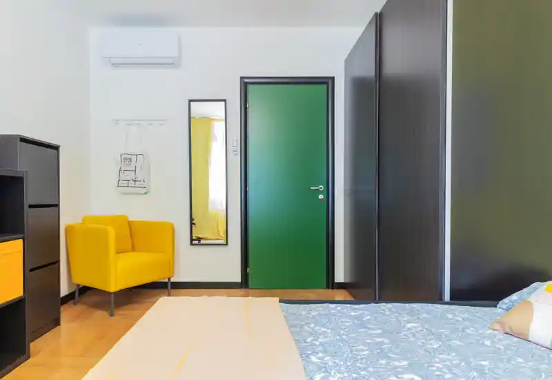 Camera da letto moderna e luminosa con letto in vista, biancheria a motivi e una poltrona gialla vivace accanto a una porta verde.