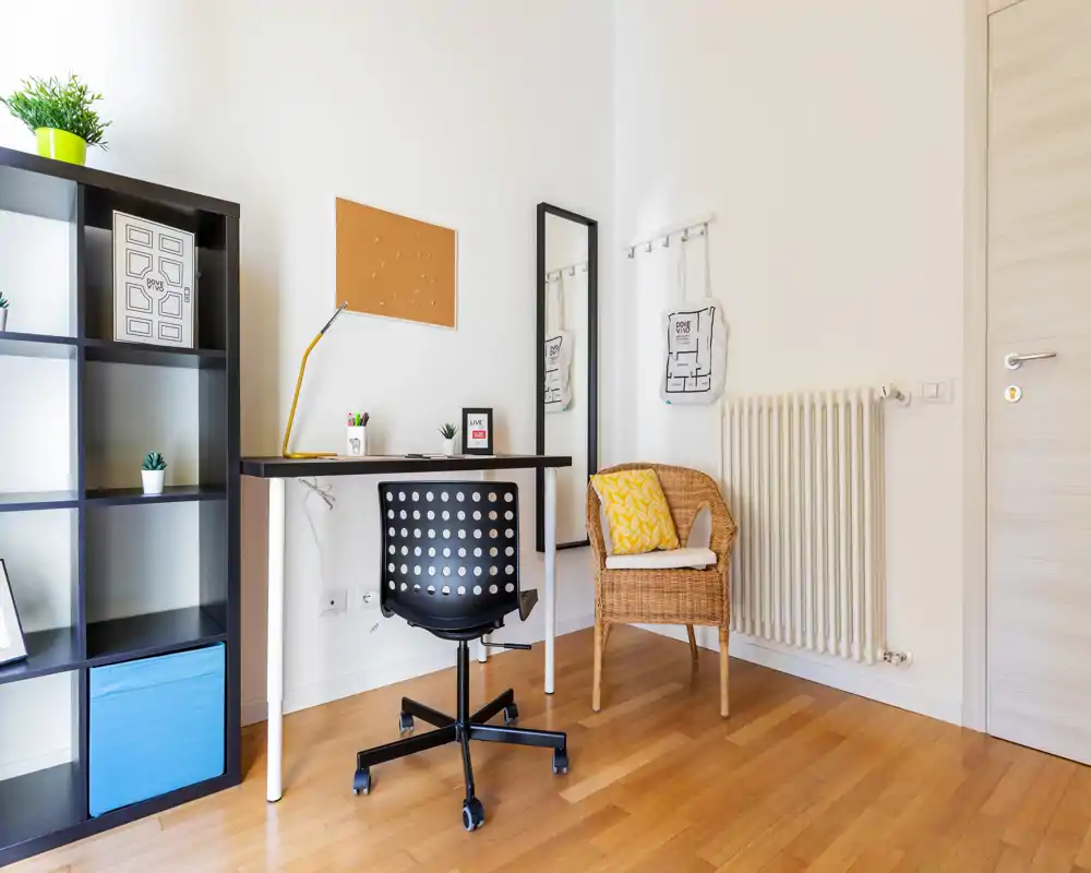 Rincón luminoso y ordenado para trabajo/descanso con escritorio, silla giratoria y butaca de mimbre — ideal como oficina en casa o zona de estar pequeña. Líneas limpias, suelo de madera cálido y detalles decorativos sencillos dan una sensación acogedora.