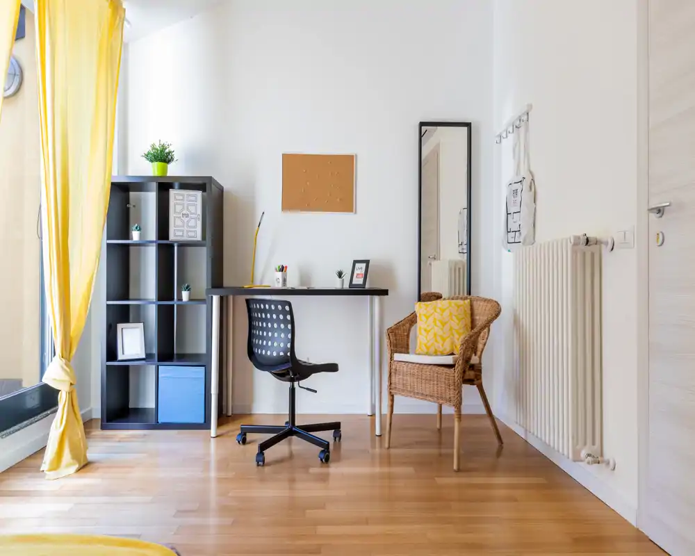 Área de estar luminosa y moderna con un pequeño escritorio, estantería y un sillón de mimbre; una ventana soleada con cortinas amarillas aporta calidez.