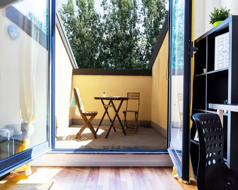 Balcón soleado con pequeña mesa bistró de madera y dos sillas plegables, visto a través de puertas correderas abiertas — ideal para el café de la mañana.