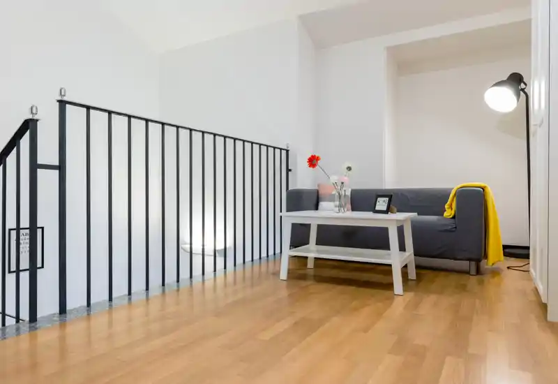 Pequeña zona de estar moderna con sofá gris, mesa de centro blanca, flores decorativas y lámpara de pie; luminosa, minimalista y bien decorada para un rincón acogedor.