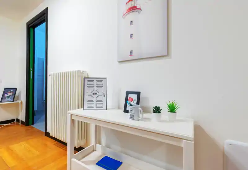 Entrada/hall luminoso y minimalista con una consola estrecha, plantas decorativas, marcos y un radiador; paredes blancas y suelo de madera cálido ofrecen una primera impresión agradable.